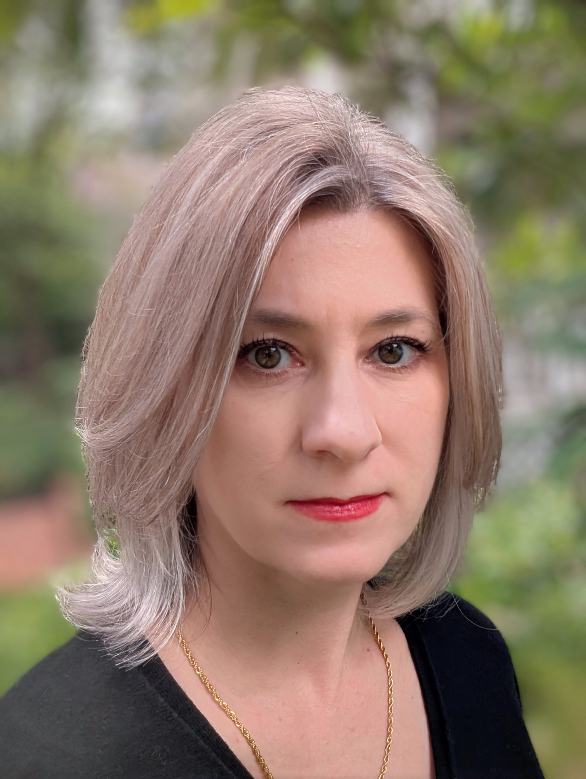 A white woman with ash blond hair in a black shirt with red lipstick. She is wearing a gold necklace standing in front of a garden in summer.